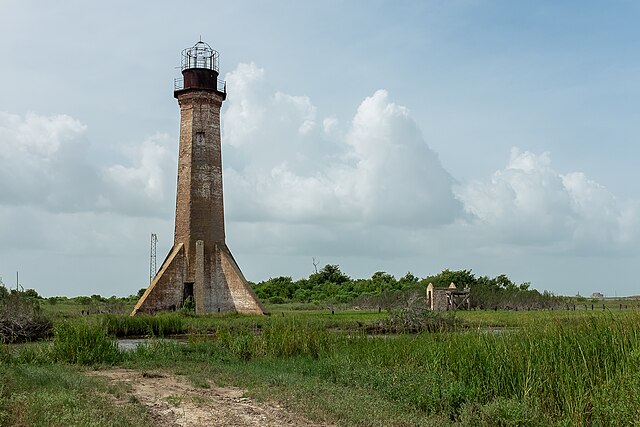 Tall, weathered brick lighthouse rising from a grassy marsh landscape under a cloudy sky, with a narrow lantern room at the top and a small abandoned structure nearby.